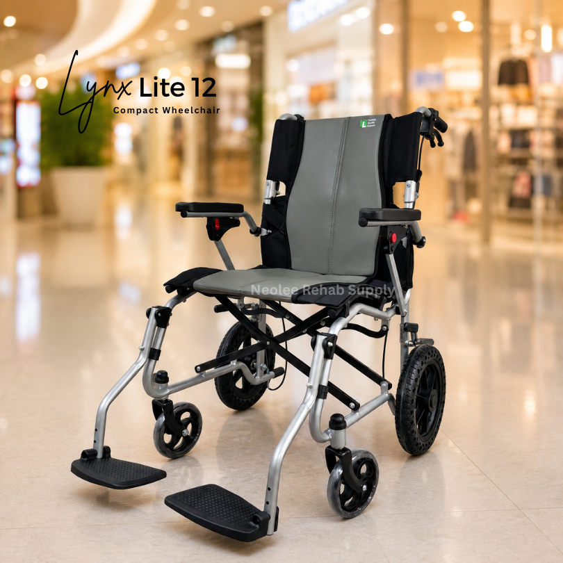 A compact silver and grey lightweight Lynx lite 12 inch wheelchair is displayed prominently in the center of a bright modern shopping mall interior with softly blurred storefronts and warm ambient lighting in the background, emphasizing mobility and accessibility.