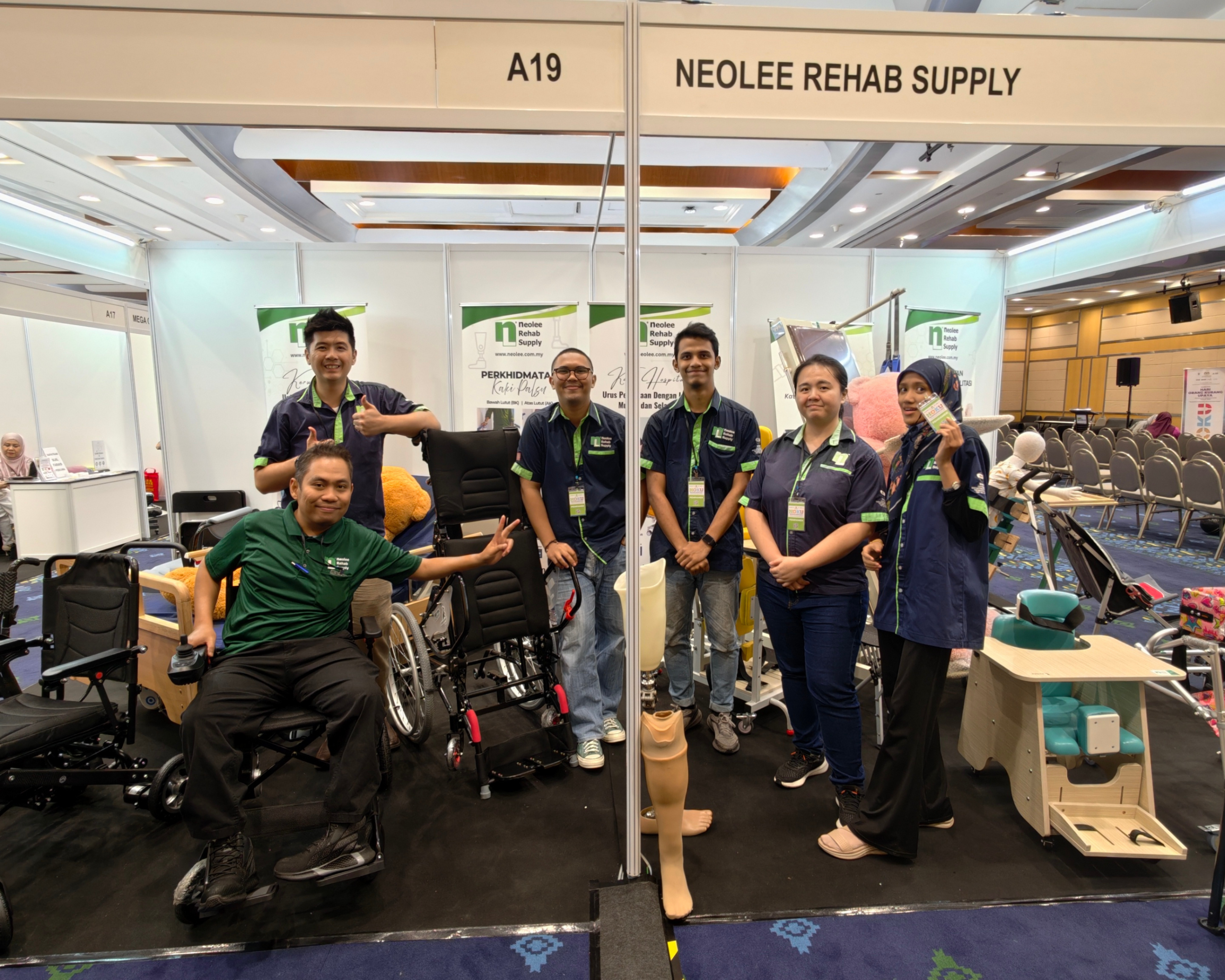 A group of five NeoLee Rehab Supply employees posing for a photo at their exhibition booth (A19). Four male and two female employee wear matching navy and green shirts. One male employee, giving a peace sign, sits in a powered wheelchair. The other five stand, some giving thumbs-up, surrounded by various models of manual and powered wheelchairs, prosthetic leg components, and booth branding.