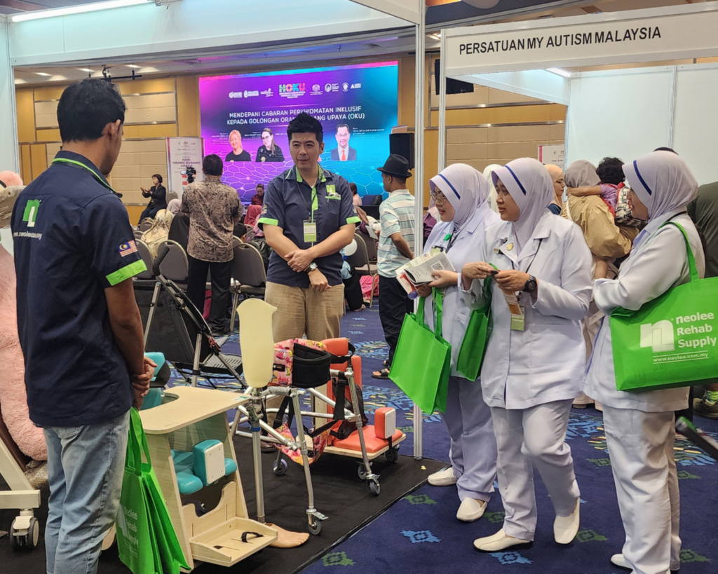 In a crowded exhibition, a NeoLee Rehab Supply representative (back to camera) and a manager speak with three women dressed as healthcare professionals or nurse. The women hold green 'NeoLee' bags. Other attendees are in the background. Prominent signs are visible, including "NEOLEE REHAB SUPPLY" branding, the main event "HOKU 2025" screen, and an adjacent "PERSATUAN MY AUTISM MALAYSIA" booth.