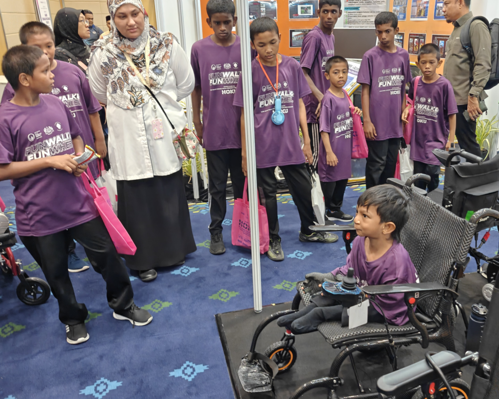 A diverse group of young participants, wearing matching purple event t-shirts that read "FUN WALK & FUN WHEEL - HOKU" (some with "OKU" on the t-shirt sleeve), are gathered at an event entrance. They are holding pink or green NeoLee bags. A woman wearing a patterned hijab is with them. Other attendees and display materials are in the background.