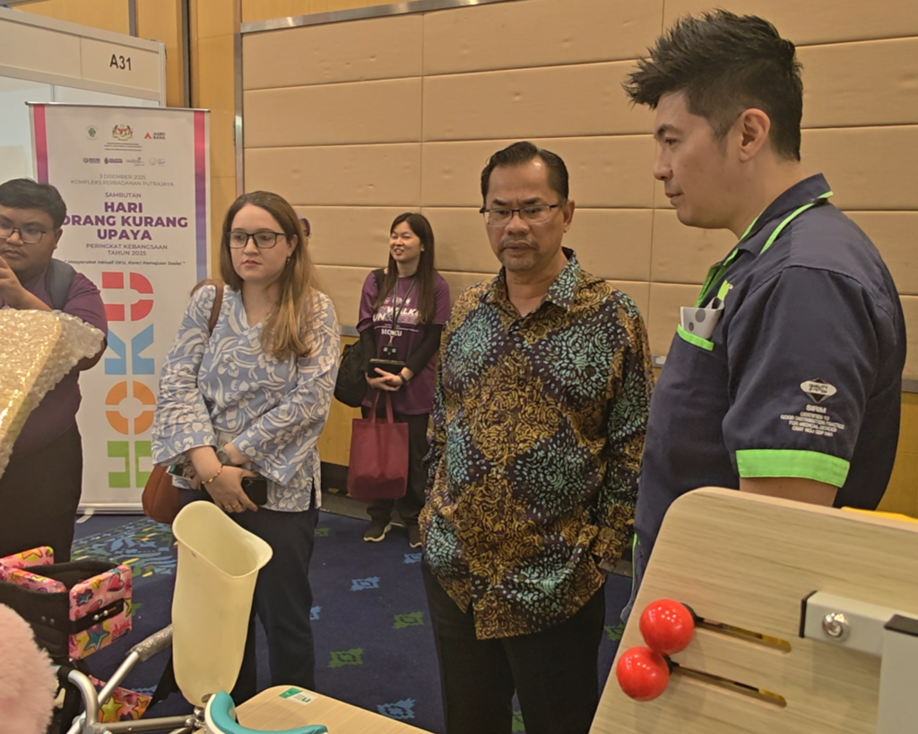 Empat orang berdiri dalam perbincangan di sebuah pameran di depan bunting untuk Sambutan Hari Orang Kurang Upaya. Seorang lelaki memakai kemeja batik biru dan kuning keemasan, dan dua wanita di sebelahnya berdiri melihat ke arah produk. Seorang lelaki memakai kemeja biru-hijau menerangkan peralatan kepada lelaki yang memakai batik. Di latar depan, terdapat peralatan pameran termasuk katil dan alat latihan fizikal untuk orang kurang upaya.