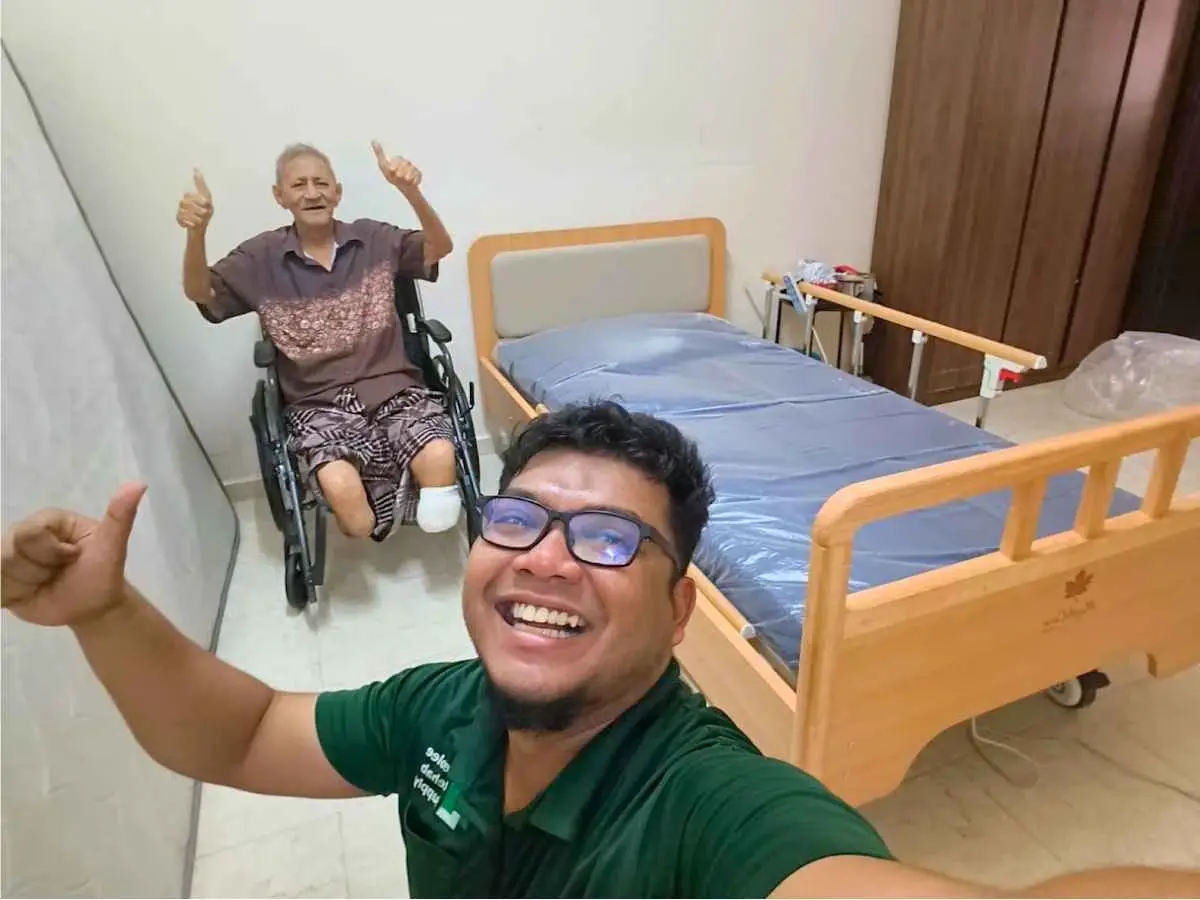 Smiling Neolee staff member taking a selfie with an elderly wheelchair user beside a Maplecare wooden hospital bed at home.